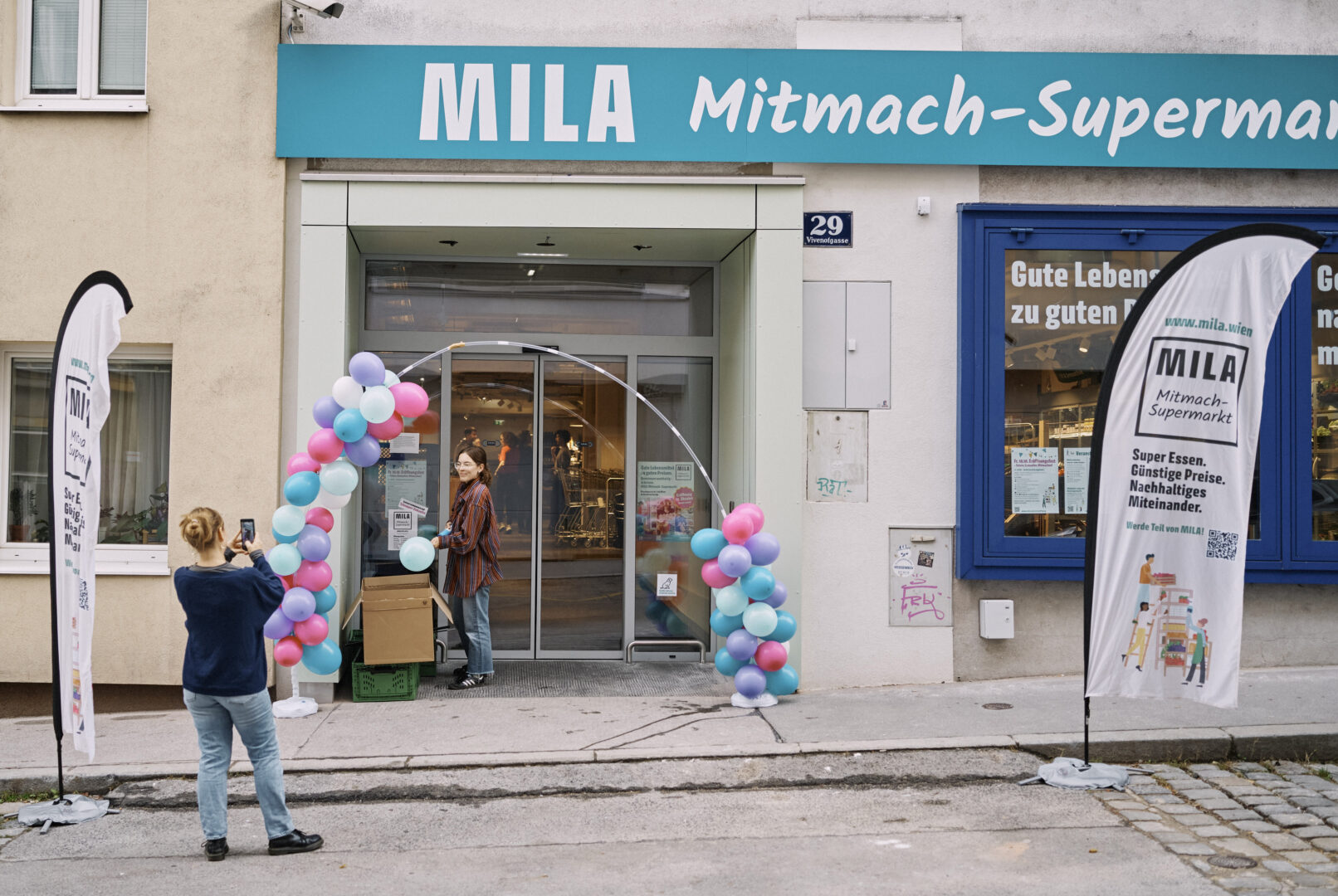 Der Eingang zum MILA Mitmach-Supermakrt am tag der Eröffnung, geschmückt mit Ballons und Beachflags. Zwei Frauen sind im Bild, die eine bei der Montage, die andere fotografiert.