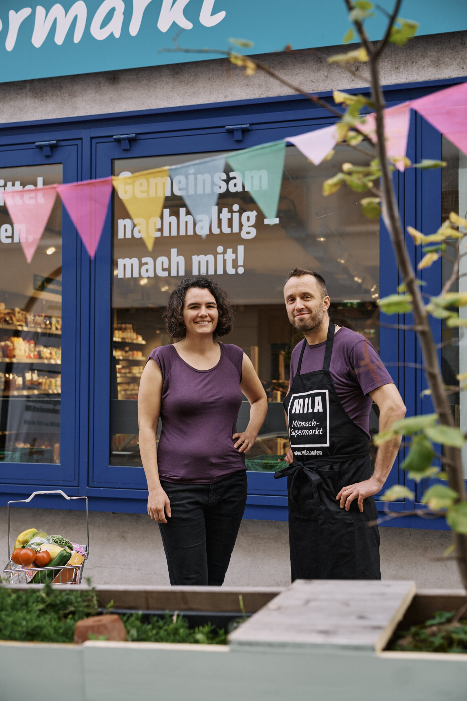 Eine Frau und ein Mann stehen vor der Auslage des Geschäftes MILA in Wien-Meidling. Beide tragen lila T-Shirts und lächeln in die Kamera. Der Mann hat außerdem eine schwarze MILA-Schürze an.