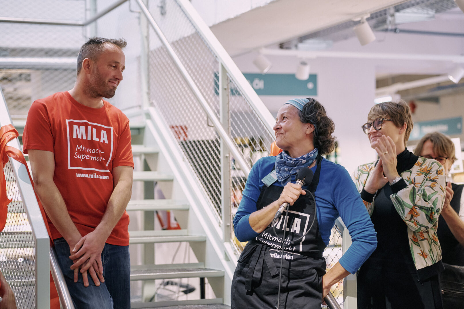 Ein Mann in MILA-T-Shirt und eine Frau in MILA-Schürze stehen an der Treppe und unterhalten sich. Die Frau hat ein Mikro in der Hand.