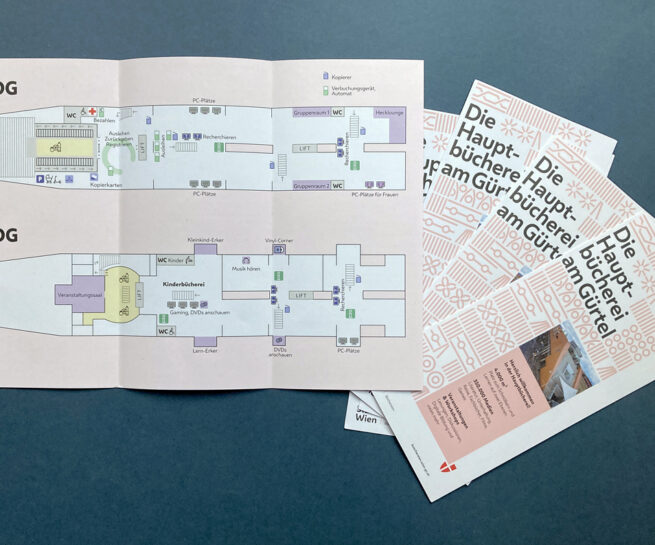 Ein Folder der Hauptbücherei Wien am Gürtel liegt aufgeschlagen dar und zeigt den Grundriß des 2. und 3. Obergeschosses. Daneben liegen mehrere geschlossene Folder, sie zeigen die Hauptbücherei am Coverbild.